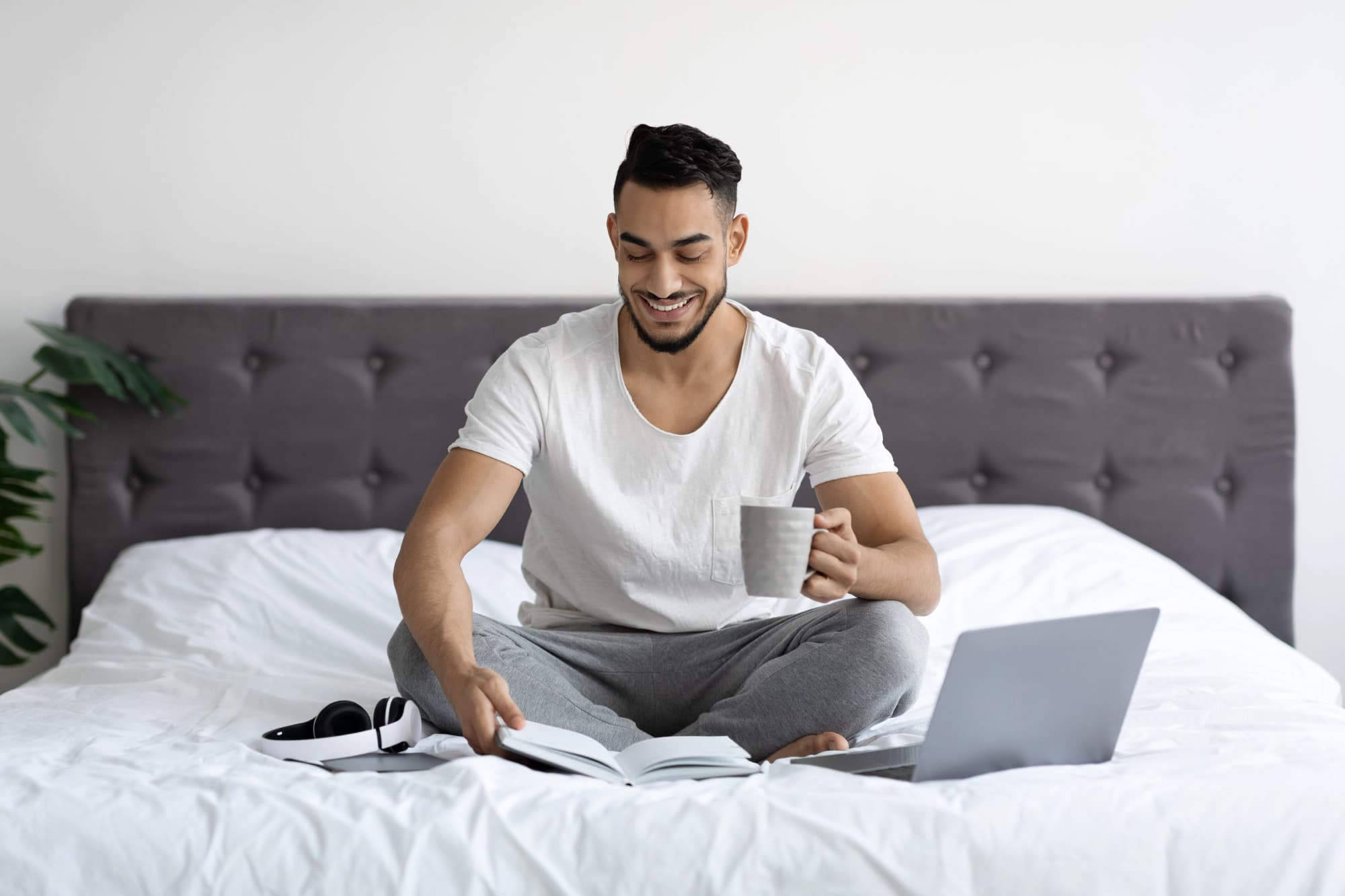 Young man sitting in bed with laptop, notebooks, headphones, and coffee.