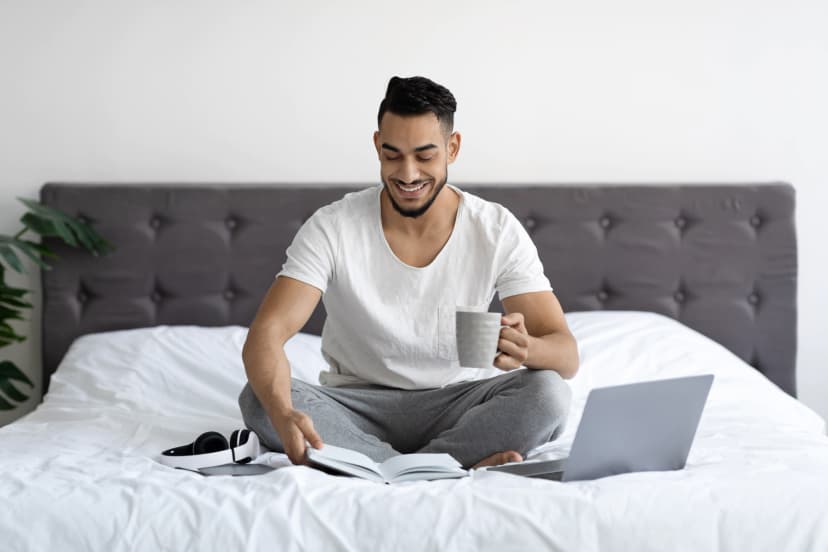Young man sitting in bed with laptop, notebooks, headphones, and coffee.