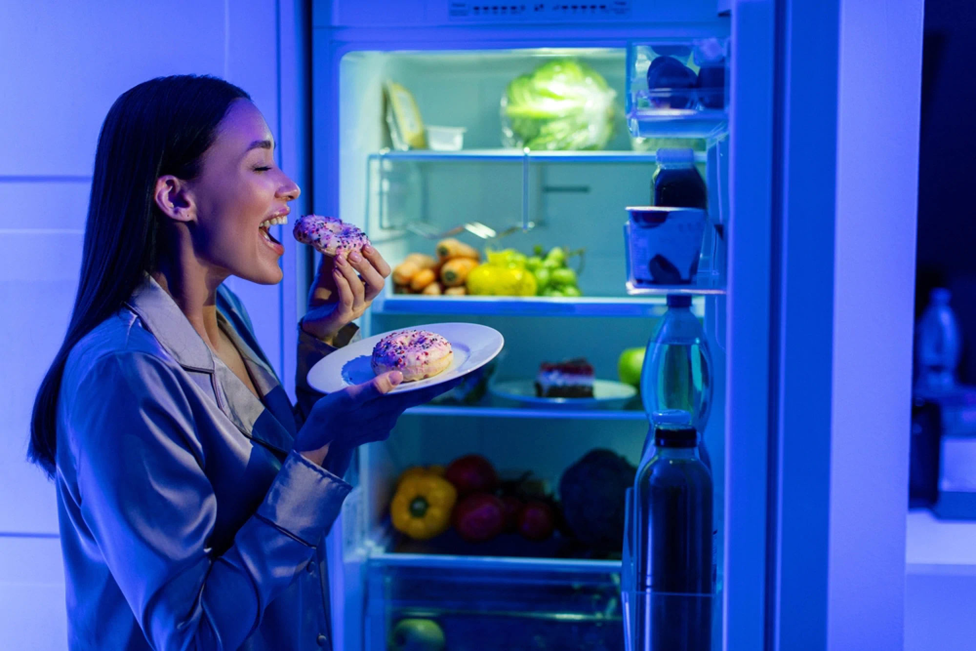 Woman in pajamas eating a donut next to her fridge