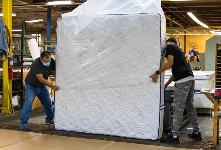 Two Texas Mattress Makers craftsmen holding up a mattress in clear plastic wrap in our factory